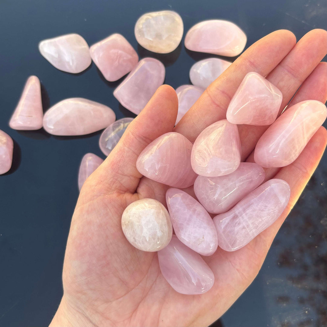 An open palm cradling a collection of mini rose quartz tumble stones, showing smooth surfaces and soft pink translucent bodies for Heart Chakra love and emotional healing.