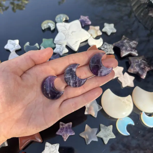 A hand holds three polished crescent moon pendants made of purple amethyst, with a collection of similar star and moon pendants scattered on a dark reflective surface in the background.