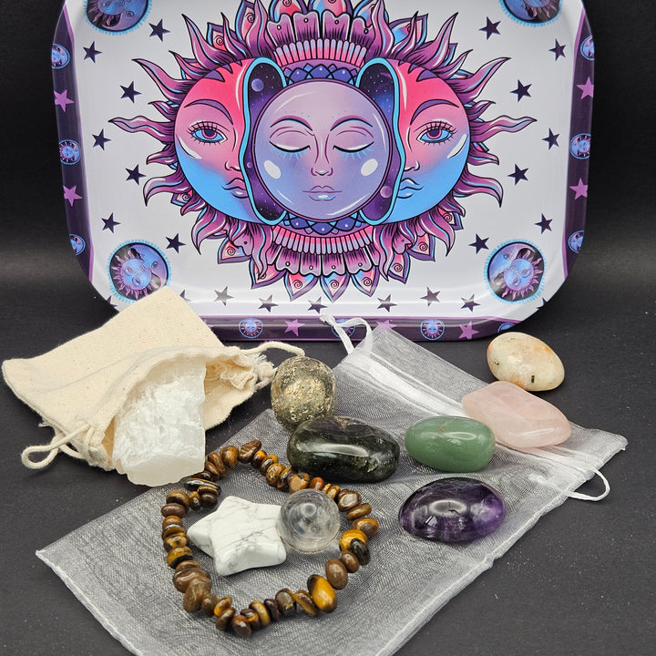 A crystal and gemstone set displayed on a dark surface with its illustrated tin box behind it. The set includes a beaded bracelet, a white star-shaped stone, a clear round stone, a pink stone, a green stone, a dark polished stone, a metallic gold-toned stone, a purple stone, a gray fabric pouch, and a cream drawstring pouch.  