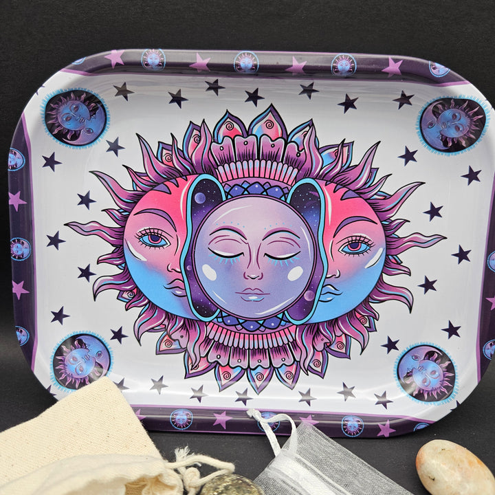 A rectangular metal tin with rounded corners, decorated with a colorful psychedelic sun and moon design in pink, purple, and blue. The tin is displayed on a dark textured surface with a small fabric pouch and stones in the foreground. 