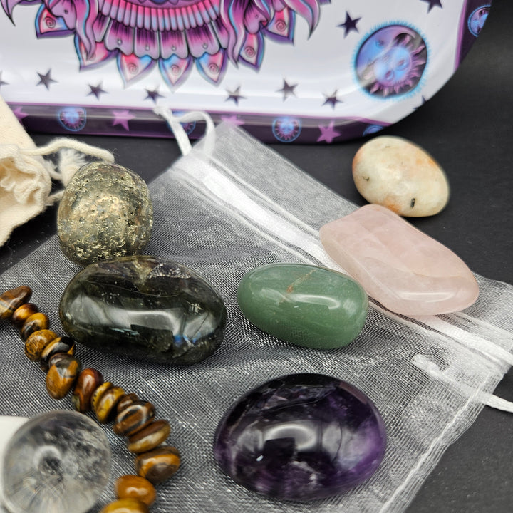 A collection of polished gemstones and crystals arranged on a white mesh drawstring pouch, including a dark green tumbled stone, a pink rose quartz chip, a purple amethyst orb, a tiger&