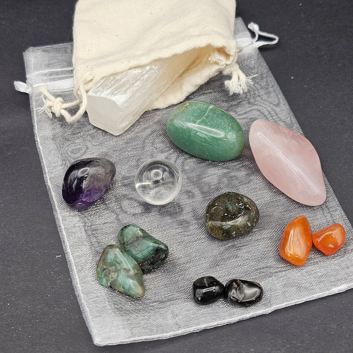 A set of polished crystal stones and a white selenite wand displayed on a gray organza pouch with a cream drawstring bag, photographed on a dark gray surface. 