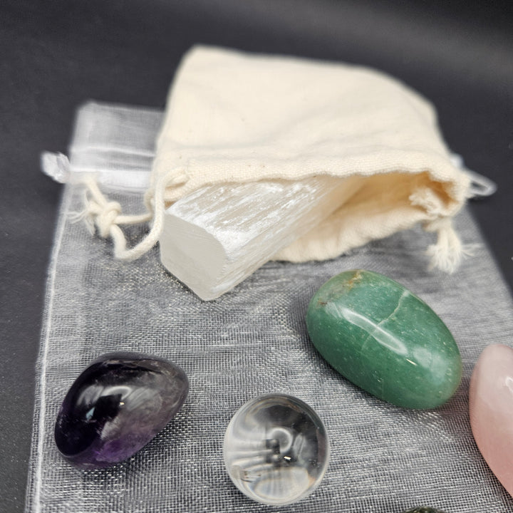 A white selenite crystal cluster wrapped in clear cellophane, resting on a gray textured cloth next to a cream drawstring pouch and several polished tumbled stones including green, purple, pink, and clear. 