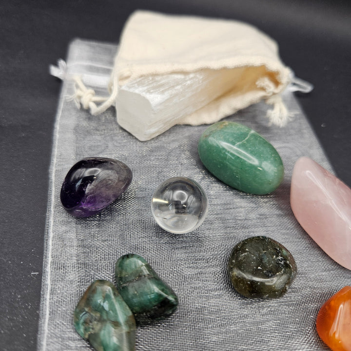 A set of polished tumbled crystals and a white selenite wand arranged on a gray textured cloth with a small cream drawstring pouch, photographed on a dark background. 