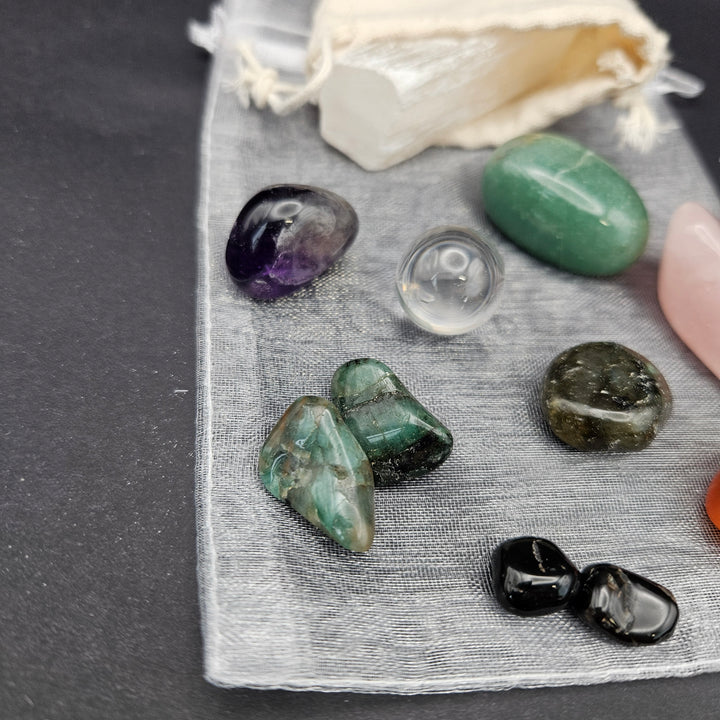 A set of polished tumbled crystals and a small cream drawstring pouch laid on a gray fabric surface against a dark gray background. Visible stones include purple, green, clear, dark green, black, pink, and orange. 