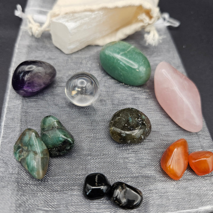 A collection of polished and tumbled gemstones in various colors, including green, pink, purple, orange, black, and clear, arranged on a gray textured cloth next to a cream drawstring pouch. 