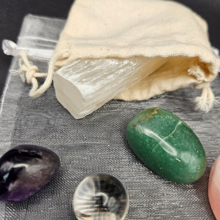 A white selenite wand resting in a small cream-colored cotton drawstring pouch, arranged on a gray textured cloth alongside polished tumbled crystals in purple, green, clear, and pink. 
