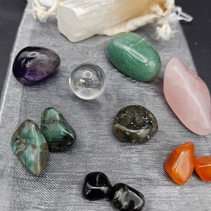 A collection of polished tumbled gemstones and crystals arranged on a gray textured fabric surface, including green, pink, purple, orange, black, and clear stones, with a small cream drawstring pouch in the background. 