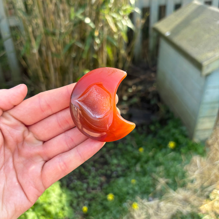 A hand holding a carved orange carnelian crystal shaped like a crescent moon against an outdoor backdrop.