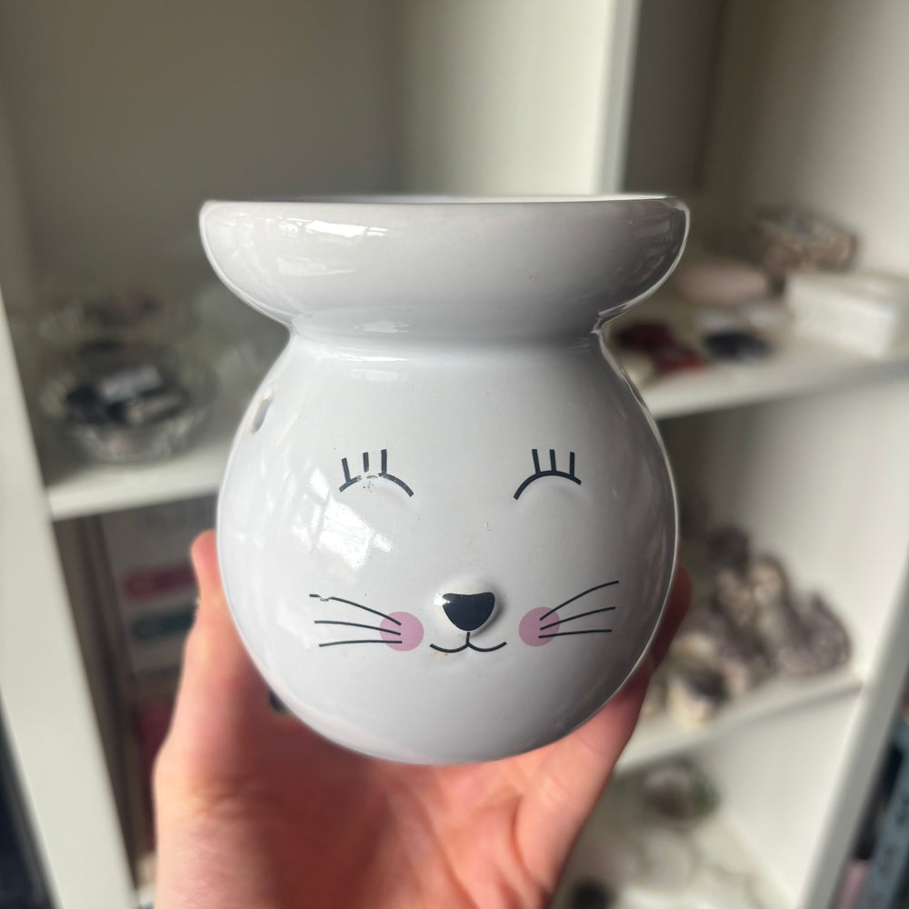 A white ceramic bunny-shaped Cat Wax Melt Burner with a cute black line-art face and pink cheeks, held in a hand against a blurred indoor background.