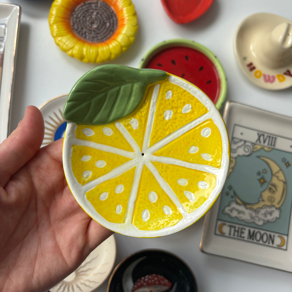 Hand holding a ceramic Lemon Incense Stick Holder with a green leaf against a background of various small ceramic items.