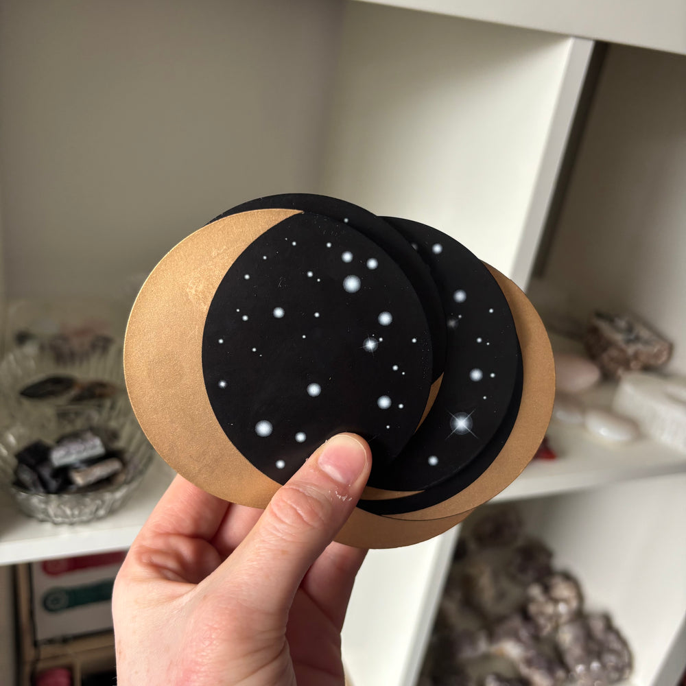 Hand holding a set of Moon Phase Coasters with black and brown design against a blurred indoor background
