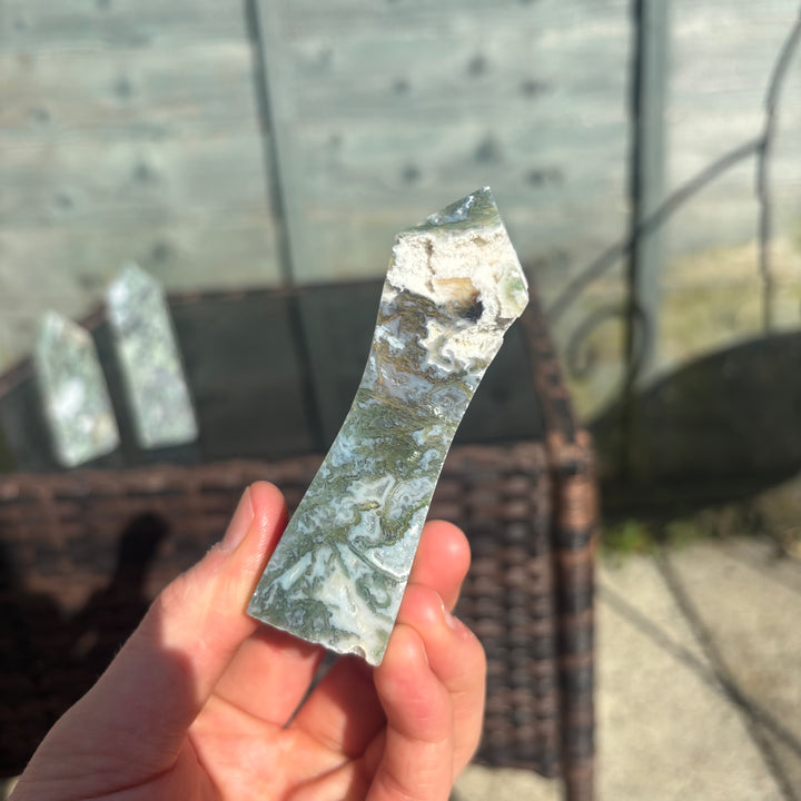 A hand holding a polished Moss Agate crystal tower with a blurred background on a bright sunny day.