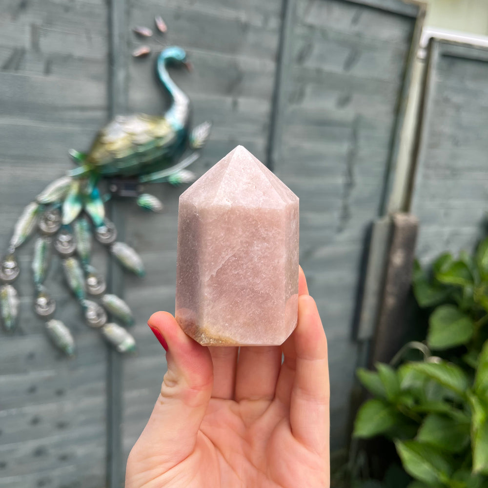 A hand holding a faceted rose quartz crystal tower against a blurred gray background with other crystals visible.