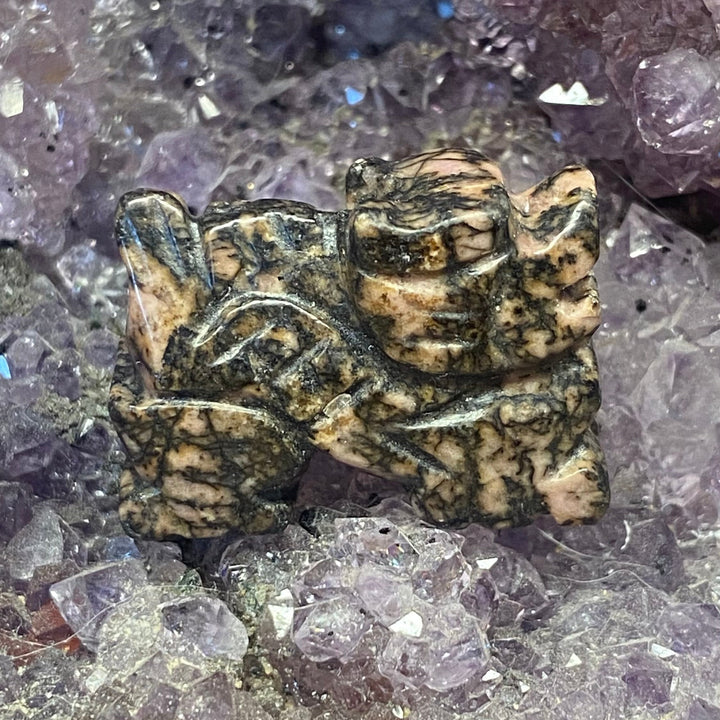 A small Rhodonite Dragon Carving with a mottled beige and dark brown pattern, resting on a bed of purple crystal clusters.