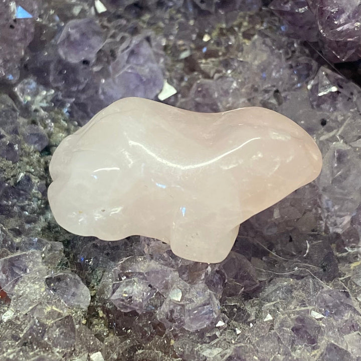 A small Rose Quartz Frog Carving, resting on a bed of purple and gray crystal clusters.