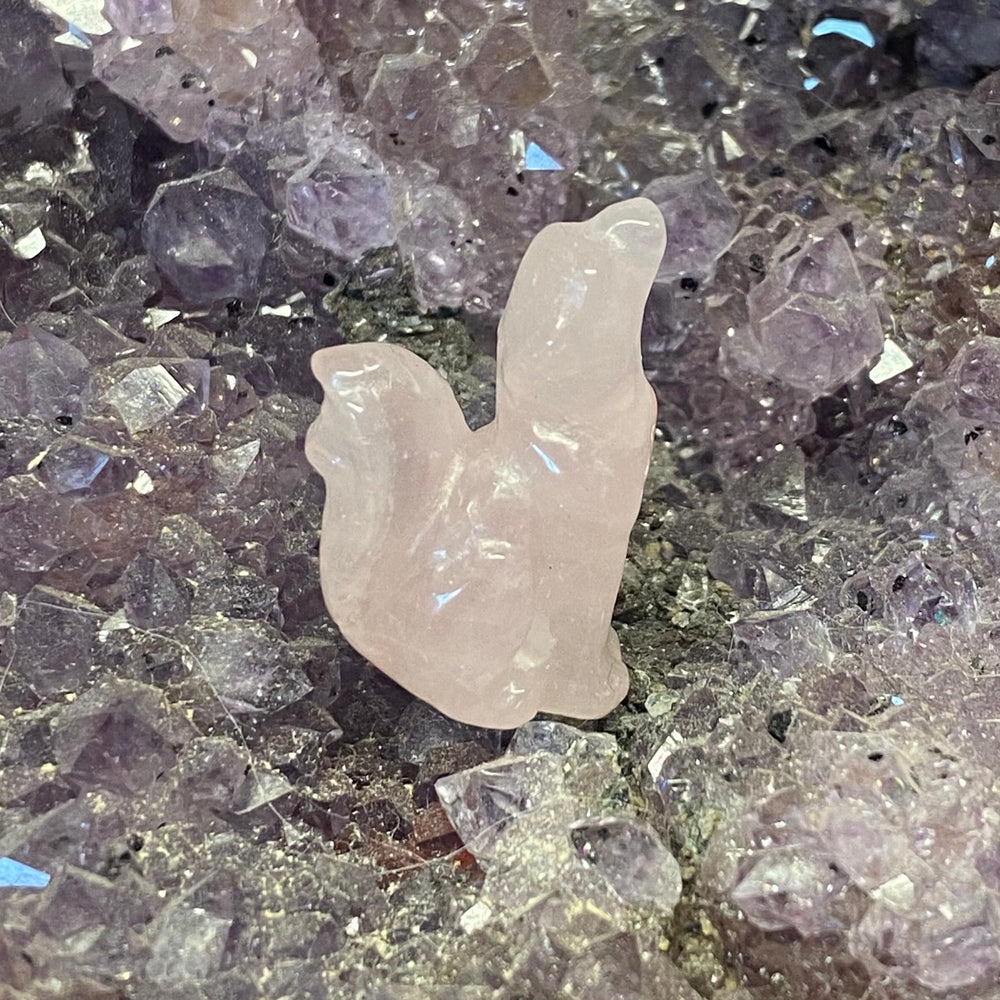 A small polished pale pinkRose Quartz Wolf Carving resting on a bed of purple and gray crystal clusters.