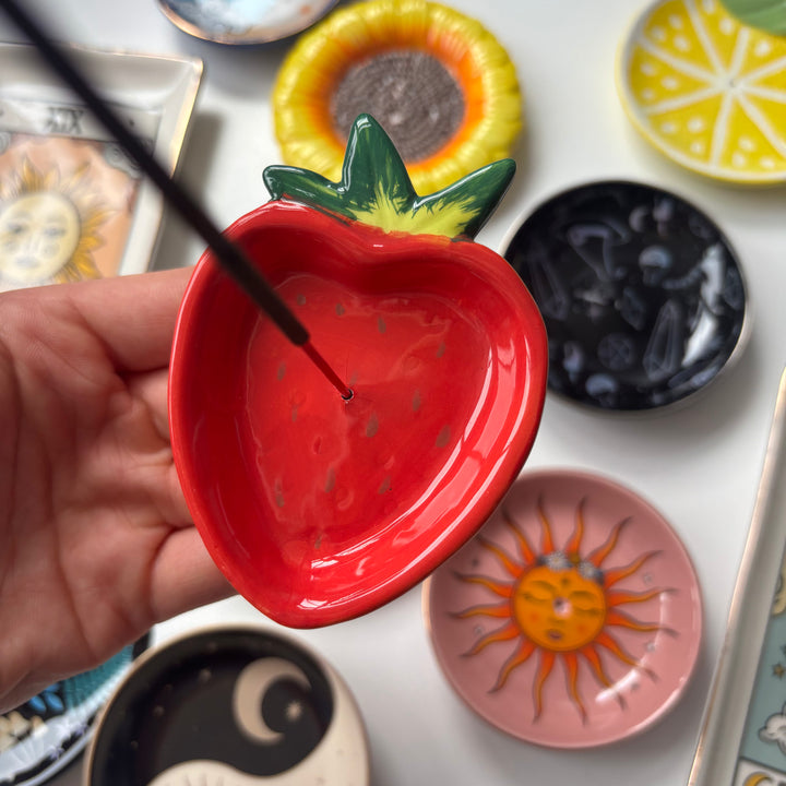 Hand holding a red Strawberry Incense Stick Holder, surrounded by various other ceramic dishes on a white surface.