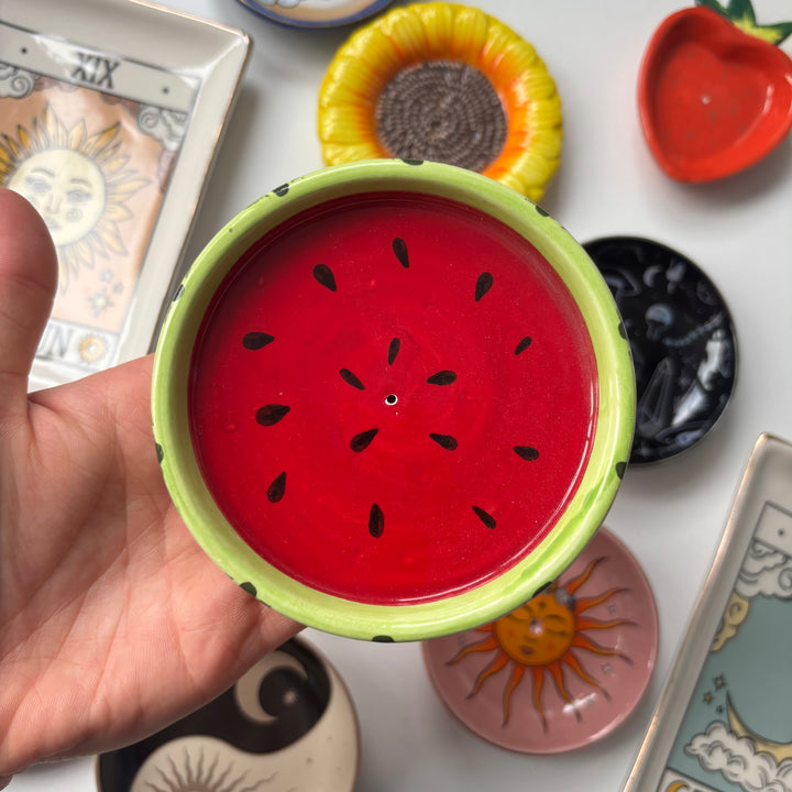 Hand holding a small red and green Watermelon Incense Stick Holder with a watermelon design against a background of various small ceramic items.