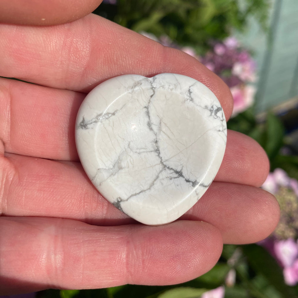 A heart-shaped Howlite crystal with gray veining, held in a person's hand with blurred pink flowers and green foliage in the background.