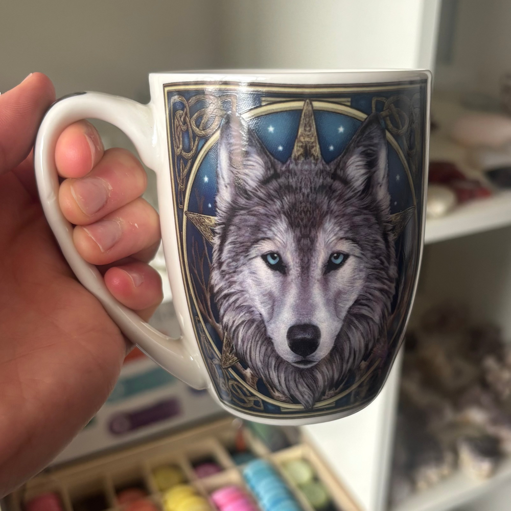 A hand holds a white ceramic mug featuring a highly detailed illustration of a gray and white wolf with blue eyes, set against a dark blue starry background with gold geometric accents.
