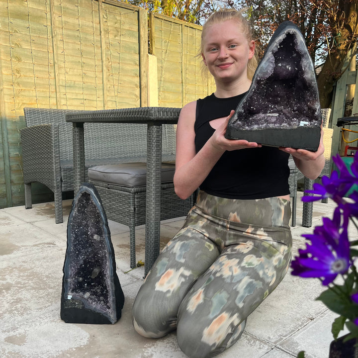 A person kneeling to display two large amethyst crystal pieces on a patio.  