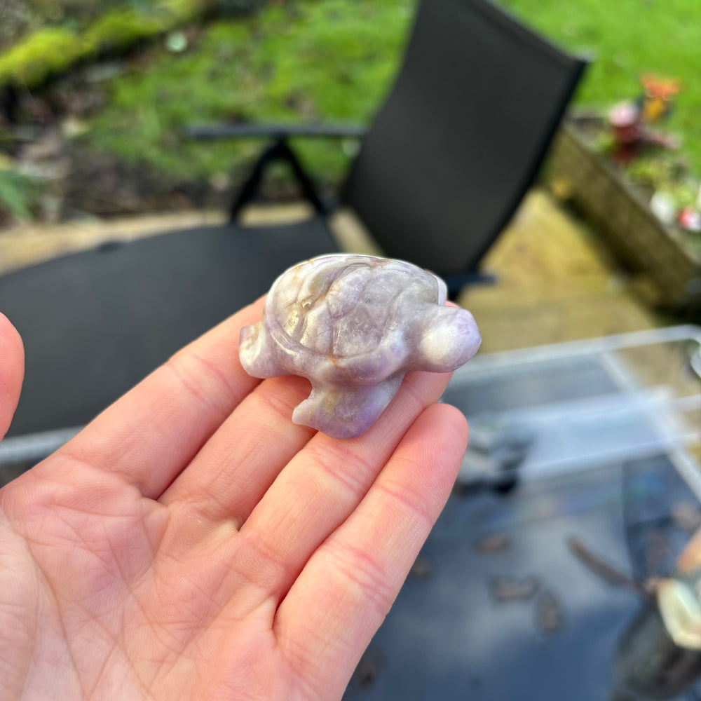 A carved amethyst turtle figurine resting on a person's fingers, showcasing its natural lavender and white crystalline texture.