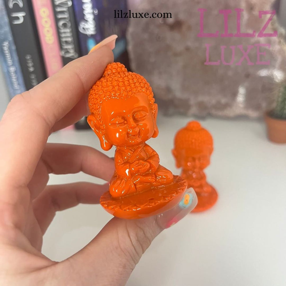 A hand holds a small Orange Baby Buddha Stick Incense Burner with a meditative pose, with another similar figurine blurred in the background.