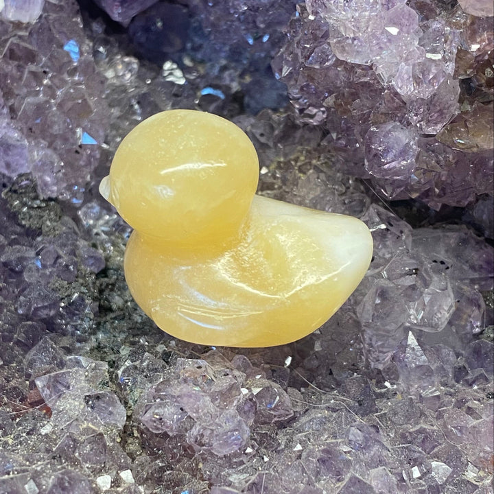 A small carved Orange Calcite Duck Carving resting on a bed of purple crystal clusters.