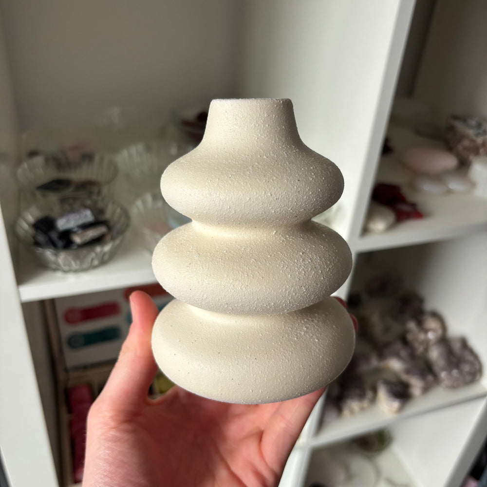 A hand holding a small, sculptural ceramic vase with a textured, matte finish. The vase features three stacked, rounded tiers with a flared top, in a warm cream or off-white color.