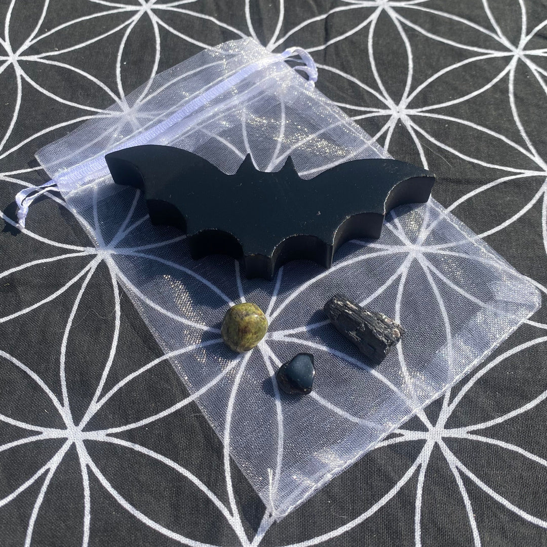 A gothic gift set arranged on a geometric Flower of Life mat, featuring a carved black wooden bat, a raw Black Tourmaline chunk, a polished Black Onyx tumbling stone, and an earthy green Dragon Stone crystal on top of a sheer white organza bag.