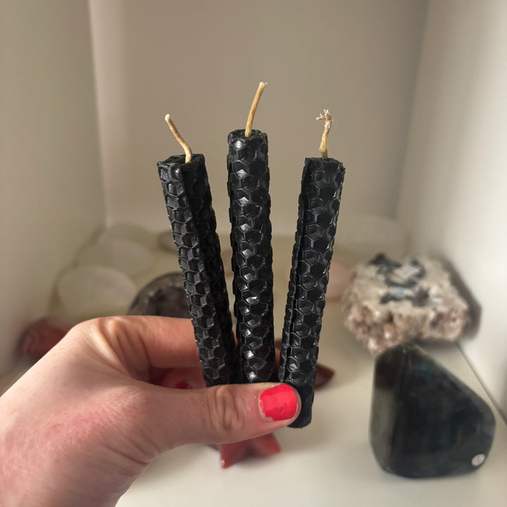 Three black textured beeswax taper candles held in a hand with bright pink nail polish, set against a softly blurred background of crystals and candles.