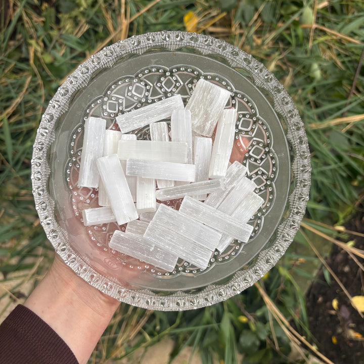 A hand holds a clear glass bowl filled with white selenite rectangular sticks outdoors, with green grass visible in the background.