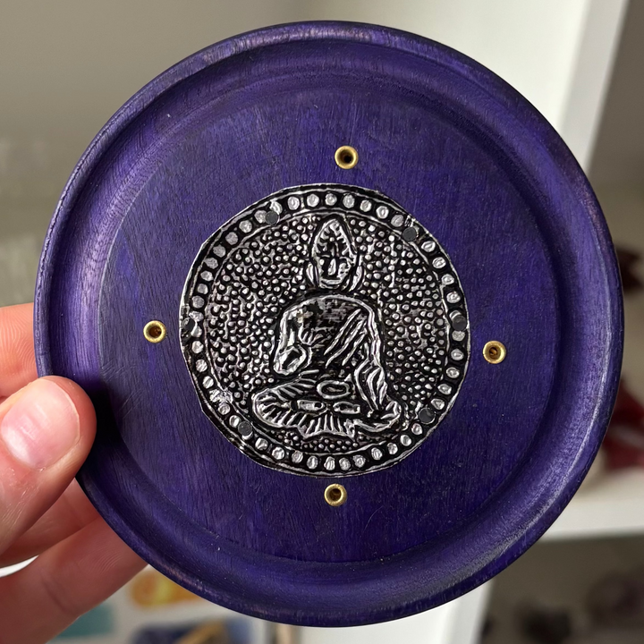 Hand holding a purple decorative item with a silver emblem against a blurred indoor background