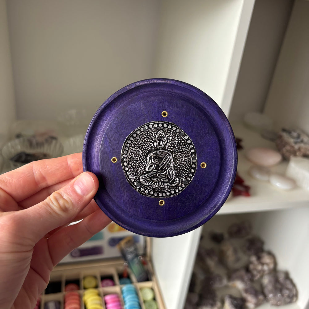A hand holding a round purple wooden plaque featuring a central metallic silver Buddha medallion and four brass screw holes around the edge.
