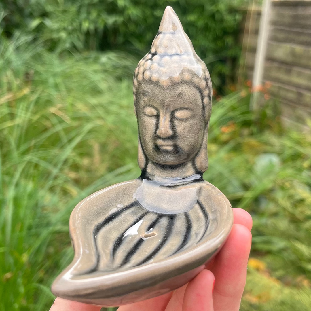 A hand holding a Buddha-shaped incense stick burner with a leaf design on top.