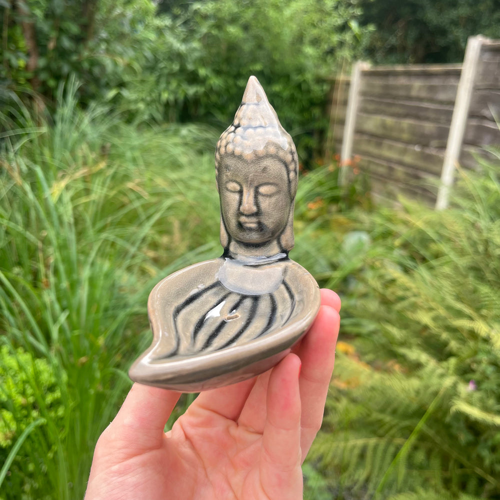 A small Buddha head incense burner with a leaf-shaped base, held in a person's hand against a blurred green garden background.