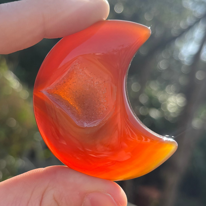 A hand holding a carved orange carnelian crystal shaped like a crescent moon against an outdoor backdrop.