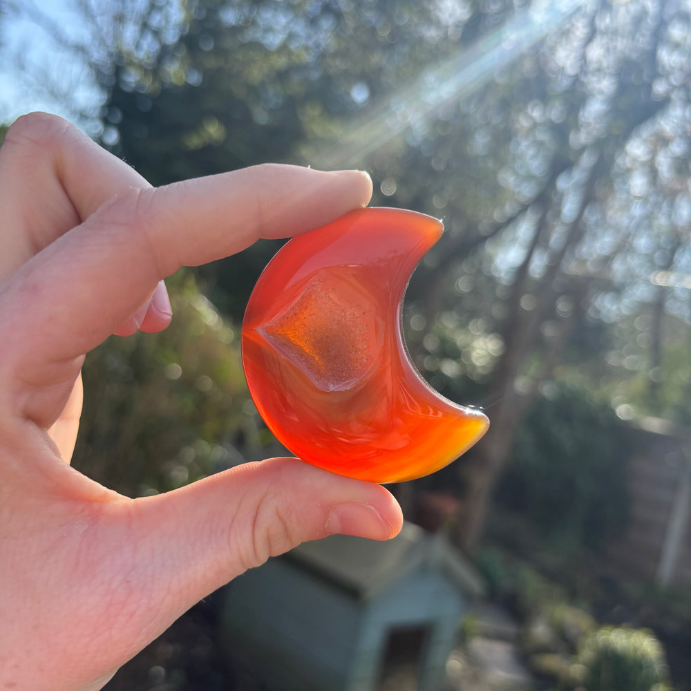 A hand holding a carved orange carnelian crystal shaped like a crescent moon against an outdoor backdrop.