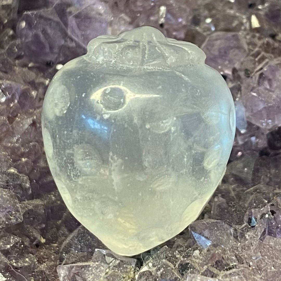 A small carved translucent Clear Quartz Strawberry Carving, resting on a bed of purple crystal points.