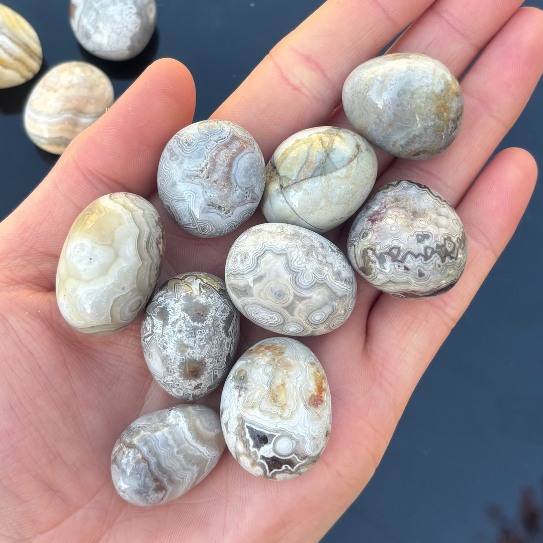 A hand holding several polished agate stone pendants with natural banded patterns in shades of gray, white, beige, and brown, set against a dark blue background.