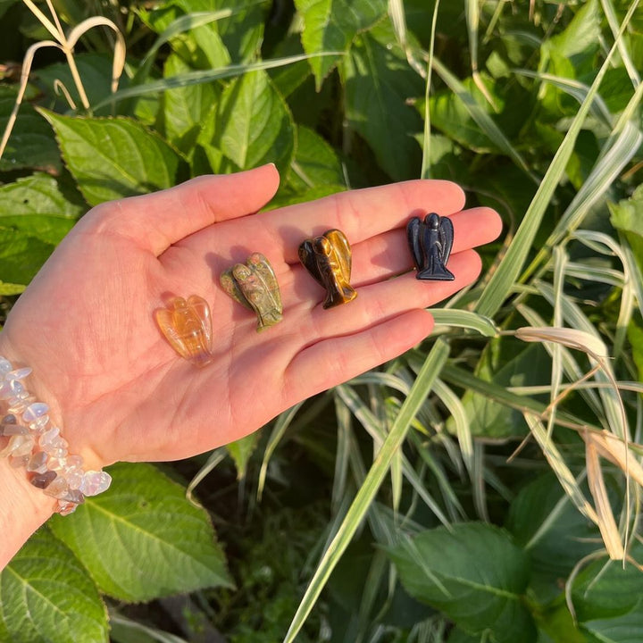 A hand holding four different colored crystal carvings of angels, with a background of green plant leaves on a bright sunny day.