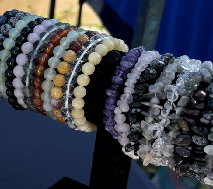 A close-up of multiple beaded and polished stone bracelets displayed on a black stand. The bracelets feature a variety of colors including purple, white, gray, black, brown, and clear stones.