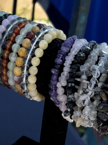 A close-up of multiple beaded and chunky crystal bracelets displayed on a black stand. The bracelets feature various stones in shades of purple, white, clear, brown, yellow, and gray, with some showing natural inclusions and polished finishes.