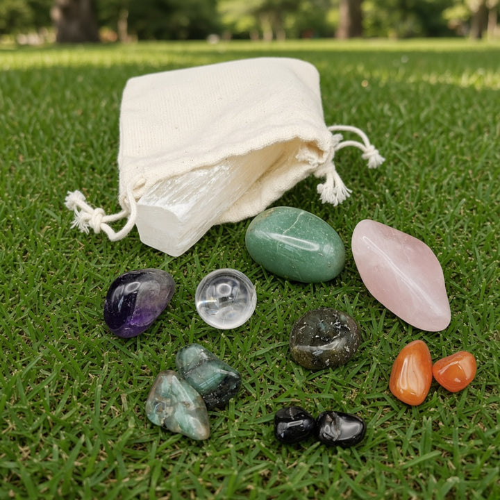 A set of polished and tumbled crystals arranged on green grass next to a small off-white drawstring pouch. The crystals include purple, clear, green, pink, black, orange, and teal stones.