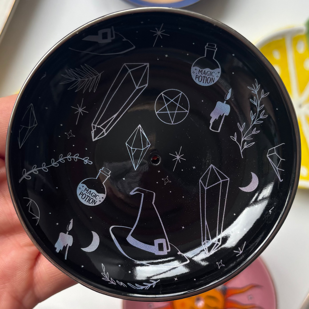 Black decorative Crystal Witch Incense Stick Holder with white designs held by a hand, surrounded by other colorful plates on a white surface.