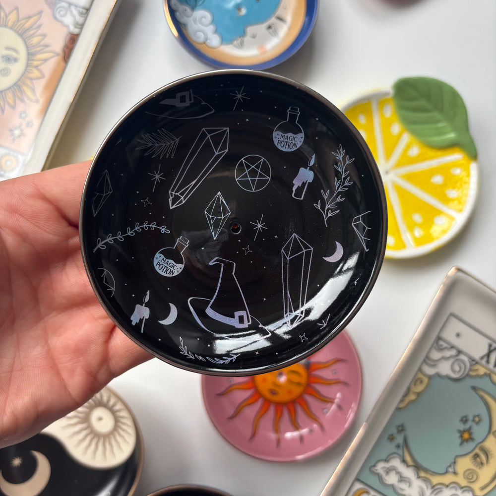 Black decorative Crystal Witch Incense Stick Holder with white designs held by a hand, surrounded by other colorful plates on a white surface.