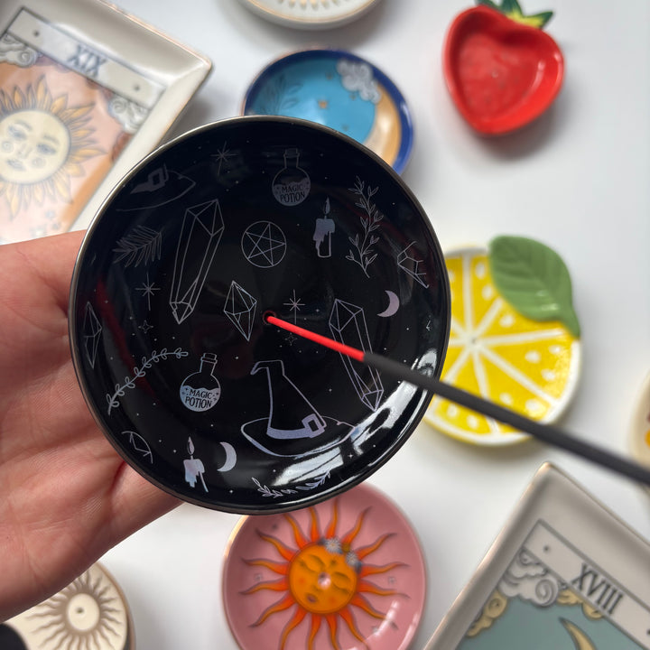 Black decorative Crystal Witch Incense Stick Holder with white designs held by a hand, surrounded by other colorful dishes on a white surface.