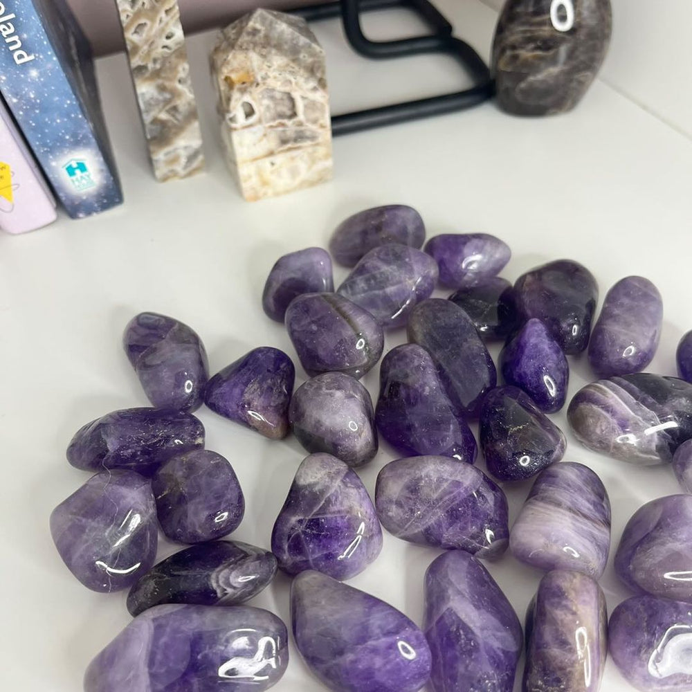 A pile of polished purple tumbled gemstones, likely amethyst, arranged on a white surface.