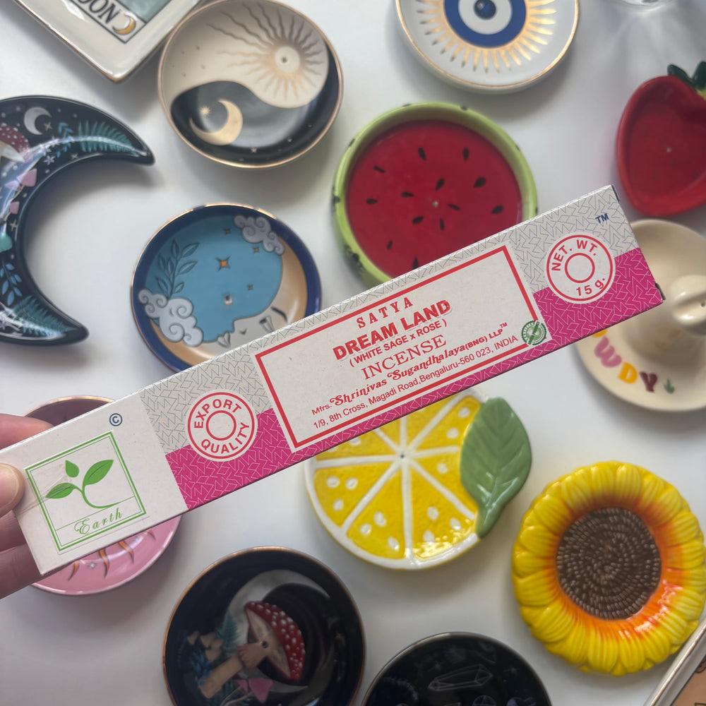 A box of Satya Dream Land Incense (White Sage + Rose) placed diagonally on a white surface surrounded by colorful ceramic dishes, including a watermelon slice, lemon, sunflower, and moon plate.